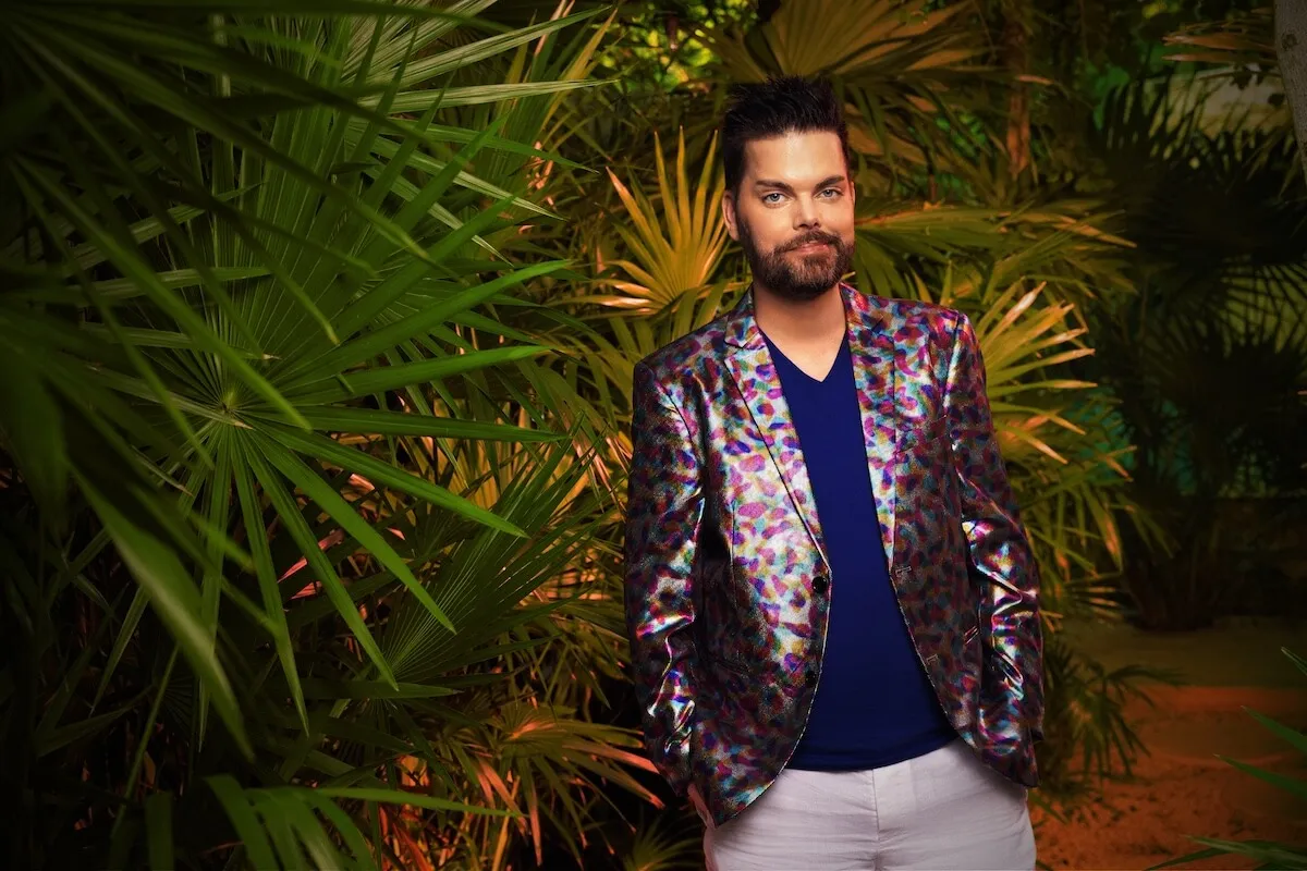 Tim Malcolm, wearing a colorful jacket, poses in front of a tropical plant in '90 Day: Hunt for Love' press photo
