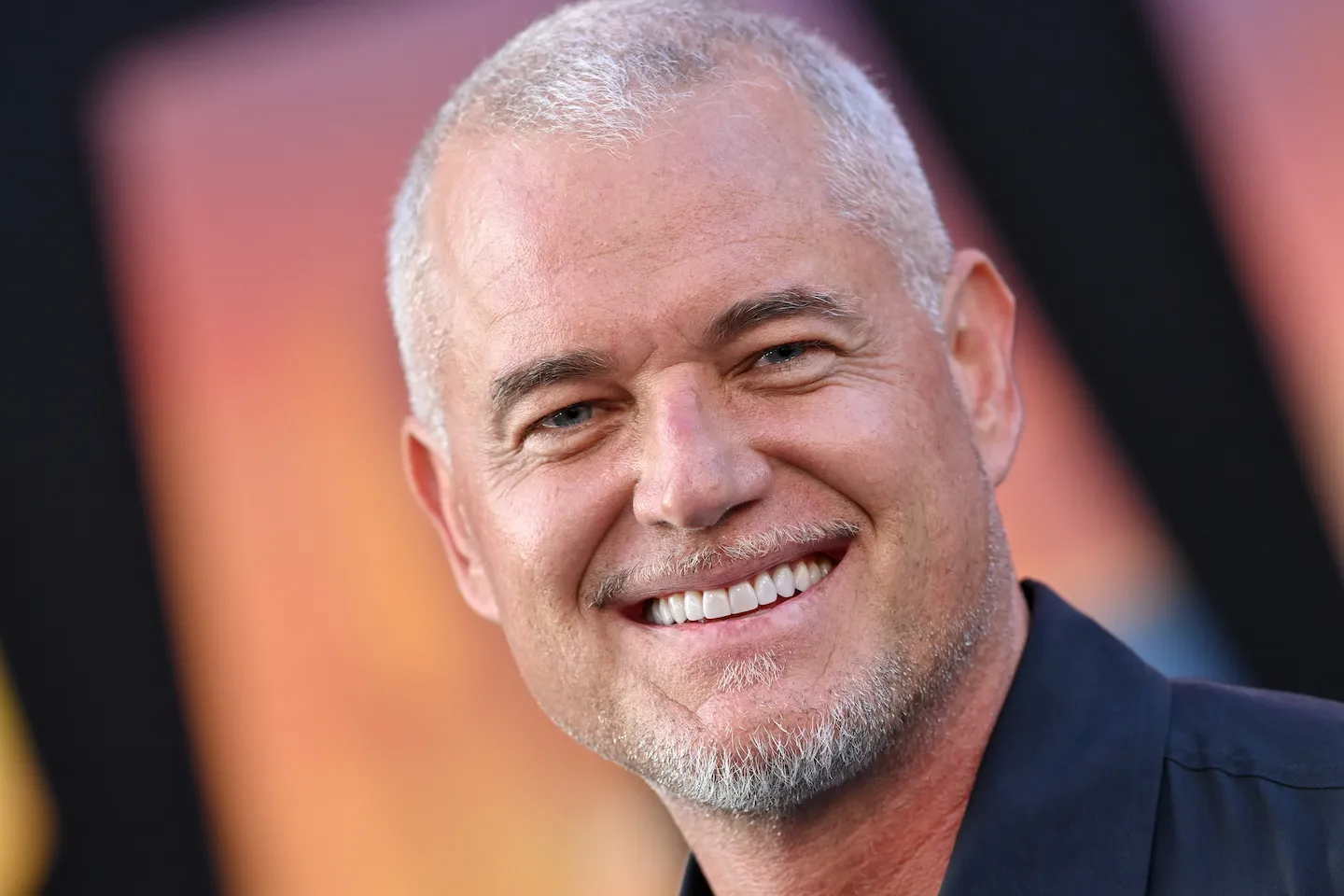 A close-up of Eric Dane smiling against an orange background