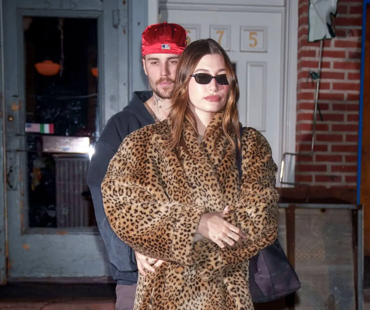 Justin Bieber wearing a red baseball cap standing behind Hailey Bieber in a leopard-print coat